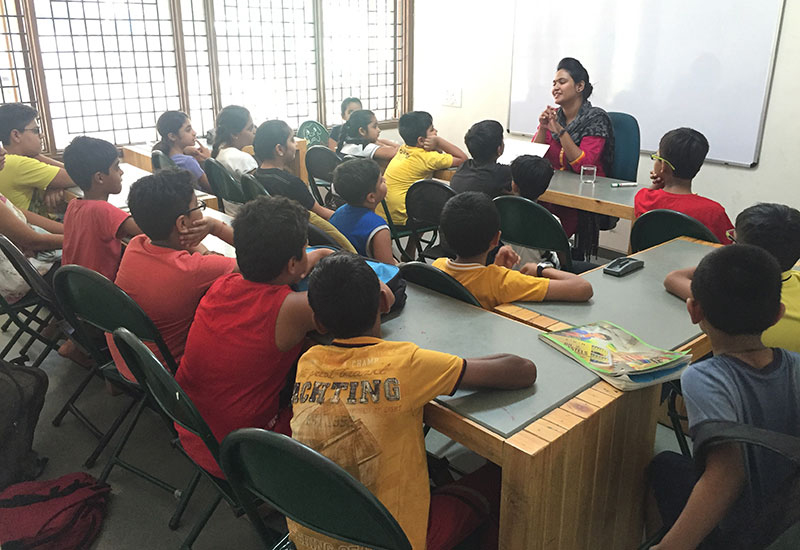 Enthusiastic group of young kids in class sitting in a row at their desk raising their hands in the air to show the know the answer to a question